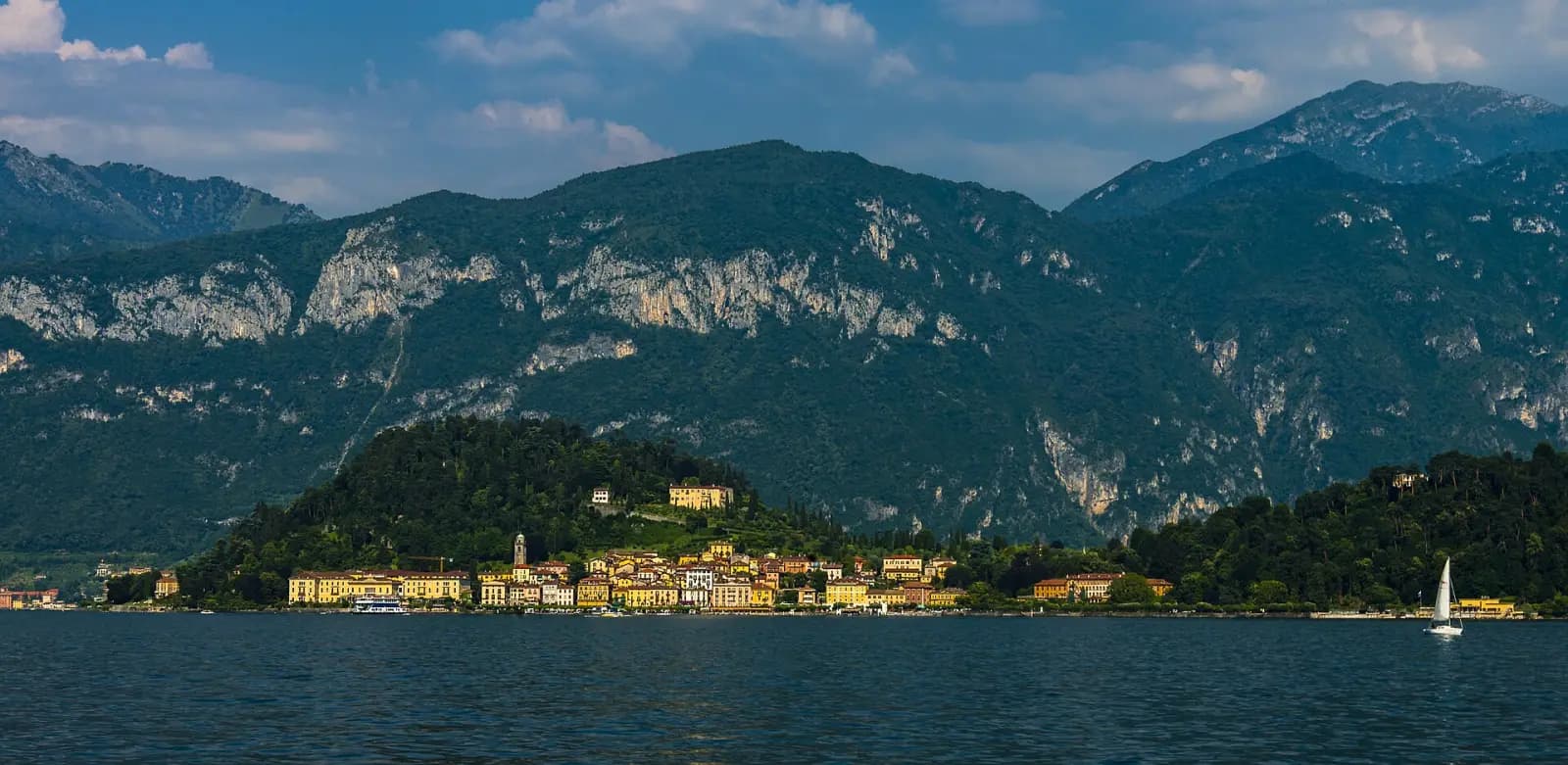 Bellagio auf seiner bewaldeten Landspitze vom See aus gesehen, dahinter Felswände der Berge