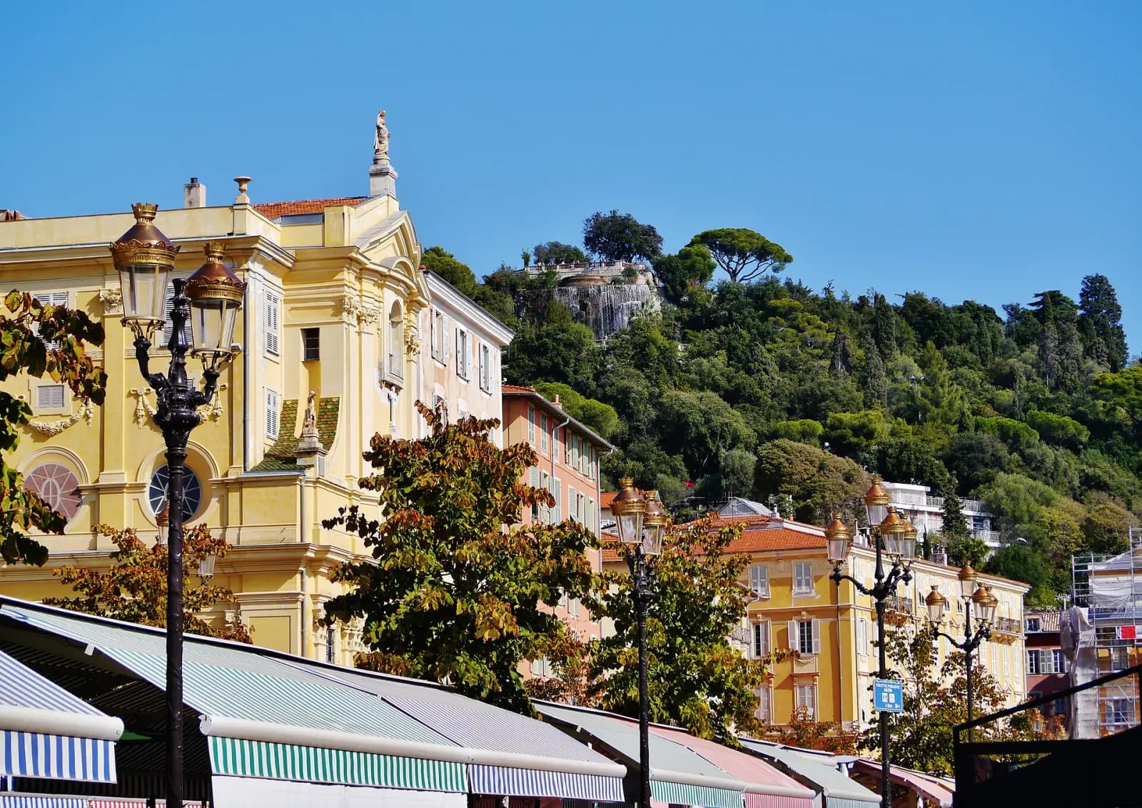 Market awnings on the Cours Saleya in front of a yellow façade, with Castle Hill behind